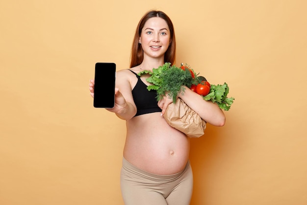 Ujmująca kobieta w ciąży trzymająca świeże warzywa pozowanie odizolowane na beżowym tle, pokazująca telefon komórkowy z pustym wyświetlaczem puste miejsce na reklamę