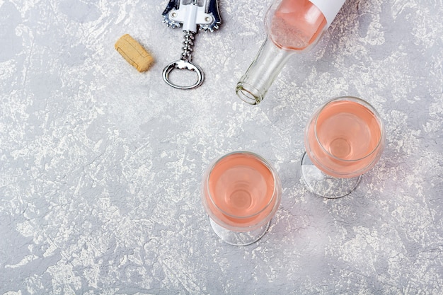 Układ do degustacji wina różanego. Otwarta butelka, dwie szklanki i korkociąg z winem różanym na szarym tle.