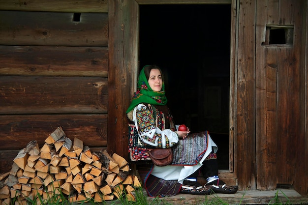 Ukrainka w narodowym stroju huculskim z jabłkiem w dłoniach siedzi na progu starego