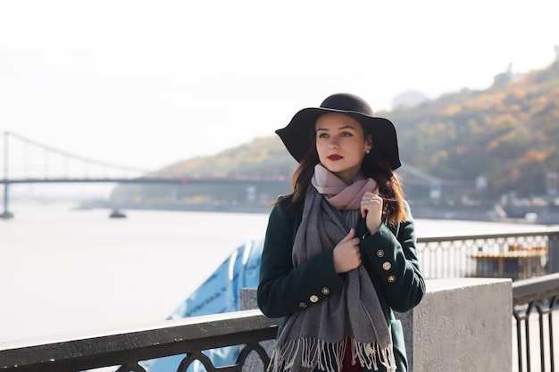 Ulica portret pięknej dziewczyny brunetka na sobie płaszcz i kapelusz i pozowanie na tle rzeki i mostu. Miejsce na tekst