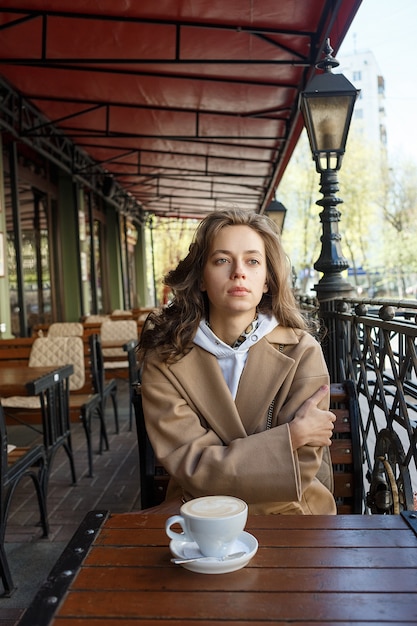Uliczny portret młodej kobiety na sobie beżowy płaszcz pije kawę na kawiarnianej werandzie z marzycielskim i zamyślonym spojrzeniem