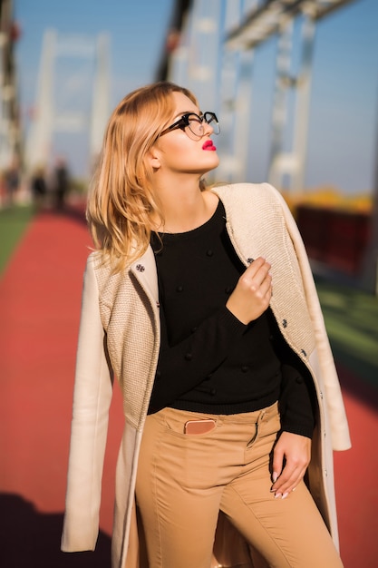 Uliczny portret uwodzicielskiej blond modelki nosi beżowy płaszcz i okulary, pozuje na moście
