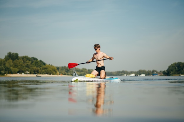 Umięśniony sportowiec trenuje pływanie po rzece na desce sup, aktywnie wiosłując z wiosłem z poważną miną