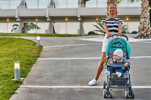 Uradowana kobieta z uroczym dzieckiem w wózku stojąca na ścieżce przed nowoczesnym hotelem w letni wieczór