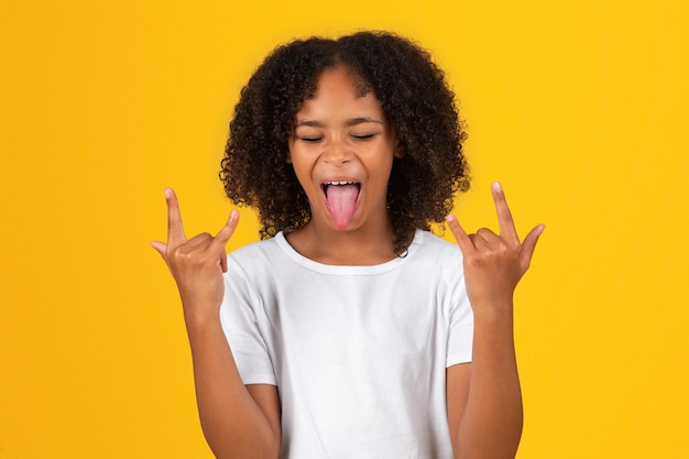 Uradowana podekscytowana nastoletnia czarna uczennica w białej koszulce pokazuje język gestem kozy rękami