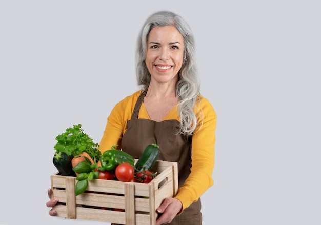 Uradowana stara europejska siwowłosa kobieta w fartuchu trzyma drewniane pudełko z organicznymi warzywami, cieszy się pracą