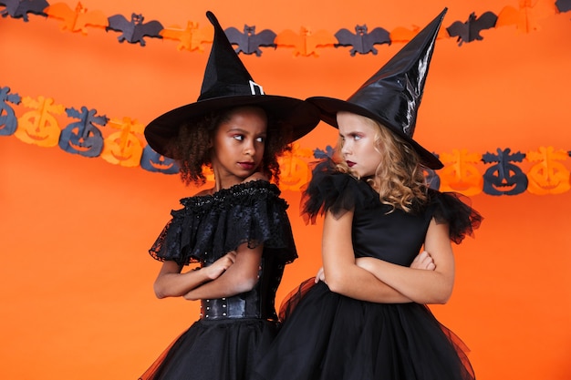 Urażone Dziewczyny Wiedźmy W Czarnym Kostiumie Na Halloween, Patrzące Na Siebie Odizolowane Nad Pomarańczową ścianą Z Dyni