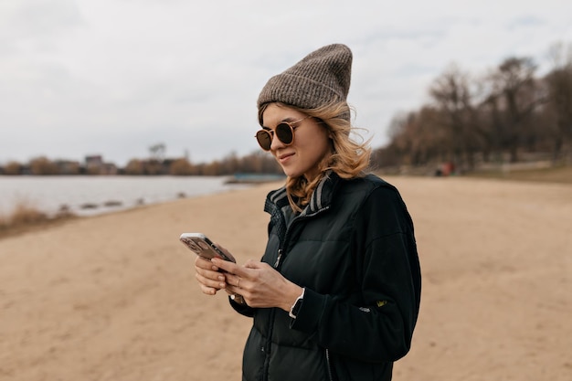 Urocza atrakcyjna kobieta w brązowej czapce i ciemnej kamizelce przewija smartfon Młoda ładna brunetka pozuje w pobliżu morza