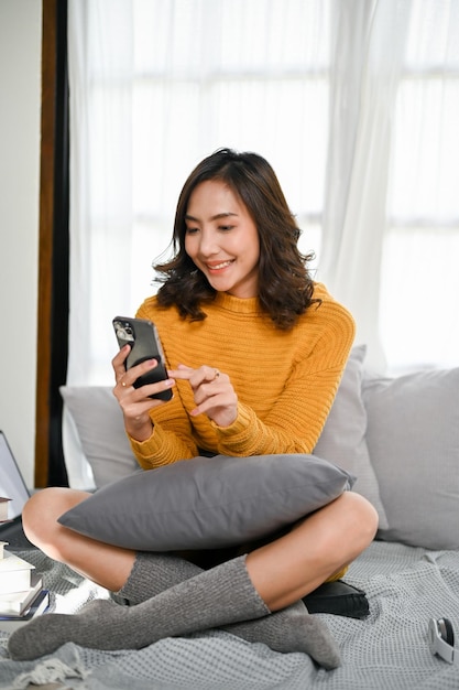 Urocza Azjatka korzystająca ze smartfona podczas relaksu na łóżku