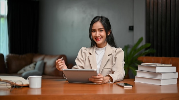 Urocza azjatycka bizneswoman używa tabletu przy biurku, uśmiechając się i patrząc w kamerę