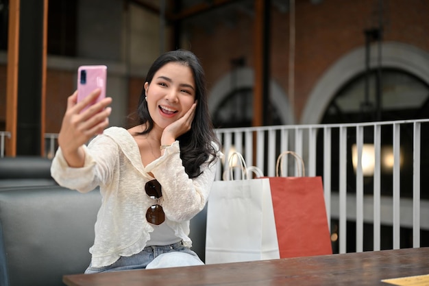 Urocza azjatycka kobieta z torbami na zakupy używająca smartfona do robienia selfie