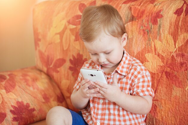 Zdjęcie urocza berbeć chłopiec obsiadanie na kanapie w żywym pokoju i bawić się z smartphone.