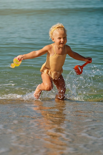 Urocza blondynka bawiąca się w wodzie morskiej na piaszczystej plaży z konewką i łopatą w rękach samotnie w ciągu dnia