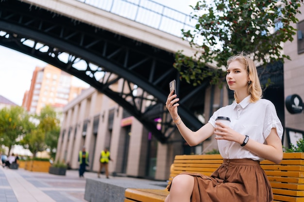 Urocza blondynka młoda kobieta bierze selfie za pomocą smartfona trzymając kawę na wynos siedząc na ławce na ulicy miasta uśmiechając się patrząc na kamerę