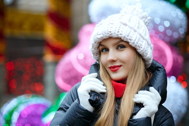 Urocza blondynka z czerwonymi ustami ciesząca się feriami zimowymi na ulicy na tle girland