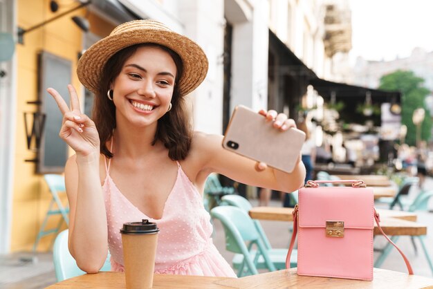 urocza brunetka kobieta uśmiecha się i bierze telefon komórkowy ze zdjęciem selfie, siedząc w letniej kawiarni na ulicy z kawą na wynos