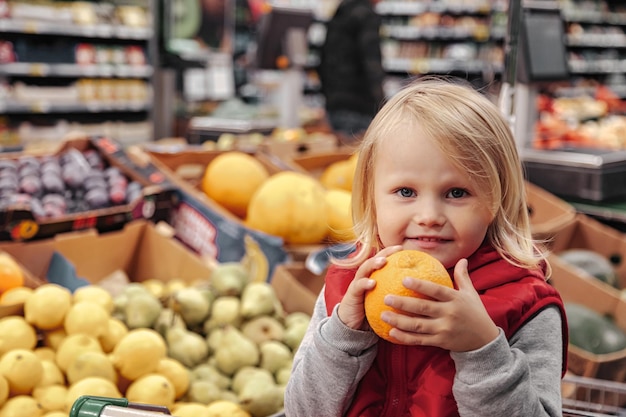 Urocza dziewczyna malucha siedzi w koszyku w sklepie z owocami żywności lub supermarkecie. Portret małe słodkie dziecko idzie na zakupy i kupowanie świeżych owoców i warzyw. Koncepcja zdrowej żywności dla dzieci