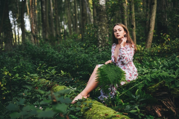 Urocza dziewczyna w kwiecistej sukni siedzi w lesie z bukietem paproci.