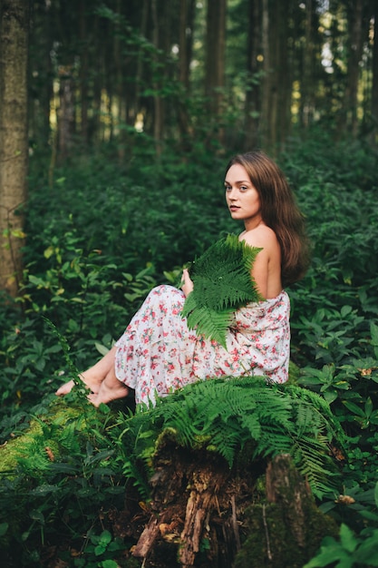 Urocza dziewczyna w kwiecistej sukni siedzi w lesie z bukietem paproci.
