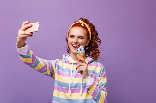Urocza dziewczyna w żółtej opasce mruga, trzyma lody i robi sobie selfie na fioletowej ścianie