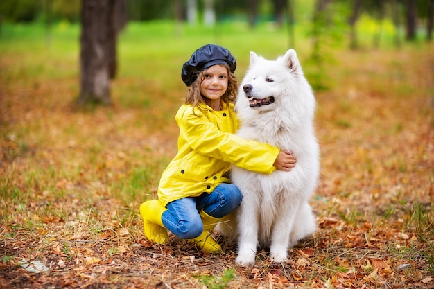 Urocza dziewczyna w żółtych kaloszach i płaszczu przeciwdeszczowym na spacerach bawi się w jesiennym parku z pięknym białym psem samojeda.