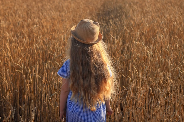 Urocza dziewczynka w słomkowym kapeluszu w niebieskiej letniej sukience na polu pszenicy Dziecko z długimi blond falującymi włosami