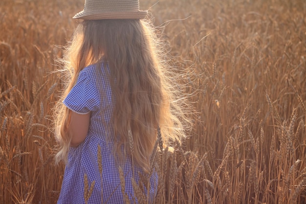 Urocza dziewczynka w słomkowym kapeluszu w niebieskiej letniej sukience na polu pszenicy Dziecko z długimi blond falującymi włosami