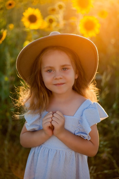 Zdjęcie urocza dziewczynka w wieku 4 lat z kręconymi długimi blond włosami w niebieskiej sukience i kapeluszu w polu słonecznika letni portret szczęśliwego dziecka zachód słońca