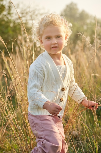 Urocza dziewczynka z kręconymi włosami, ubrana w biały sweter stojąca na zielonym trawniku w słonecznej letniej wsi i patrząca na kamerę