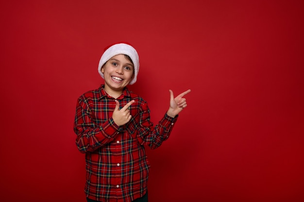 Urocza europejska preadolescent wesoły chłopiec, przystojne dziecko w Santa hat i kraciastej koszuli wskazuje palcami na miejsce na czerwonym kolorowym tle na reklamę świąteczną i noworoczną