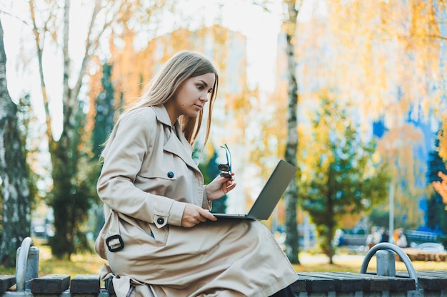 Urocza jasnowłosa młoda kobieta siedzi w jesiennym parku na ławce i pracuje nad laptopem Praca zdalna