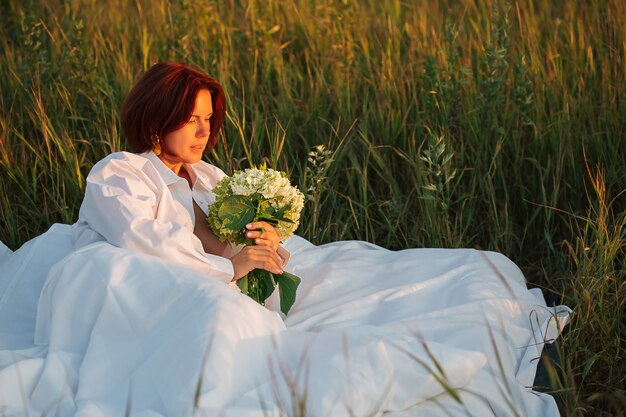 Urocza kaukaska kobieta 30-35 lat w białym łóżku w polu z bukietem hortensji w białej męskiej koszuli o zachodzie słońca latem, piękno w koncepcji natury.