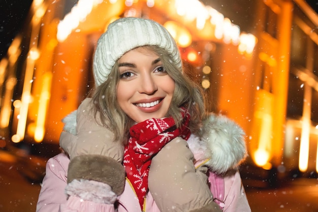 urocza kobieta na zaśnieżonej ulicy w zimowej kurtce uśmiecha się radośnie wokół wieczornych świateł
