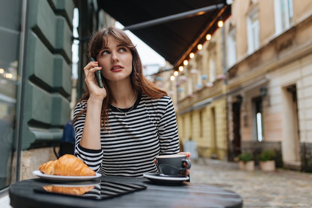 Urocza kobieta w codziennych ubraniach ma połączenie przez komórkę, delektując się świeżą kawą na tarasie kawiarni