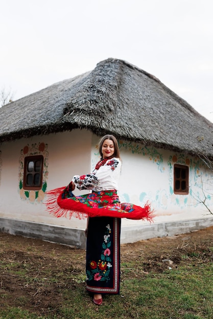 Urocza kobieta z tradycyjnym ukraińskim naszyjnikiem z chusteczką i haftowaną sukienką stojąca na tle udekorowanej chaty z dachem krytym strzechą Ludowa kultura etniczna w stylu ukraińskim