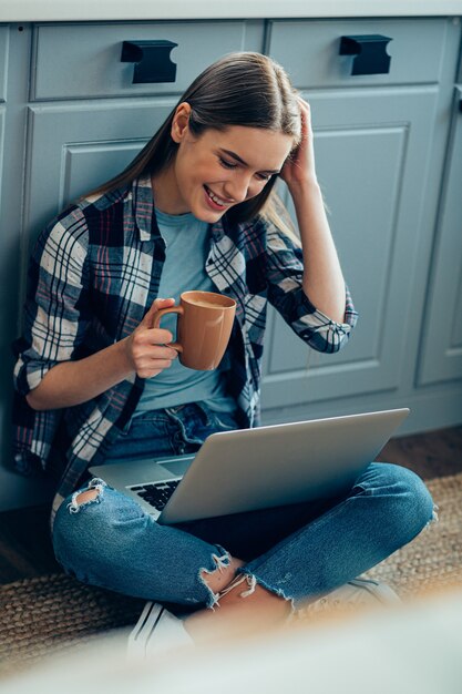 Urocza ładna Kobieta Siedzi Na Podłodze I Pije Herbatę, Patrząc Na Ekran Laptopa