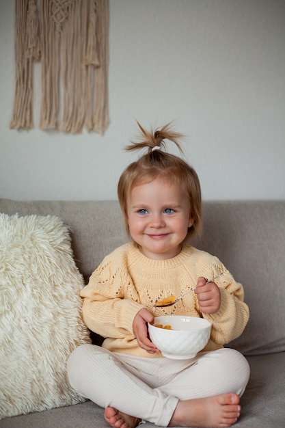 Urocza mała 2-letnia dziewczynka w przytulnym swetrze z dzianiny je śniadanie na kanapie