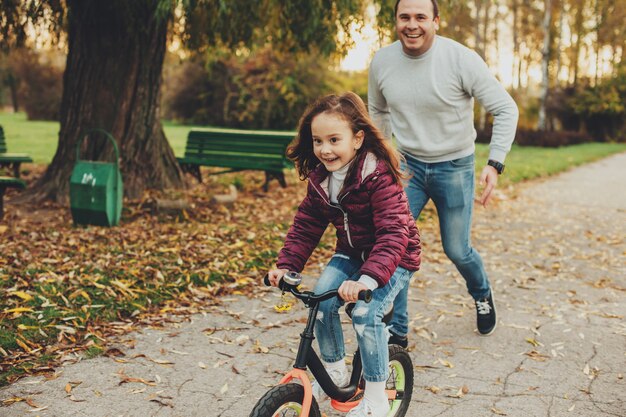 Urocza mała dziewczynka jedzie na rowerze, podczas gdy jej ojciec próbuje złapać ją na śmiechu na świeżym powietrzu w parku.