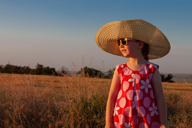 Urocza mała dziewczynka słomkowy kapelusz różowa letnia sukienka z kłoskami w ręku w widoku z tyłu pola pszenicy Długie blond włosy dziecko