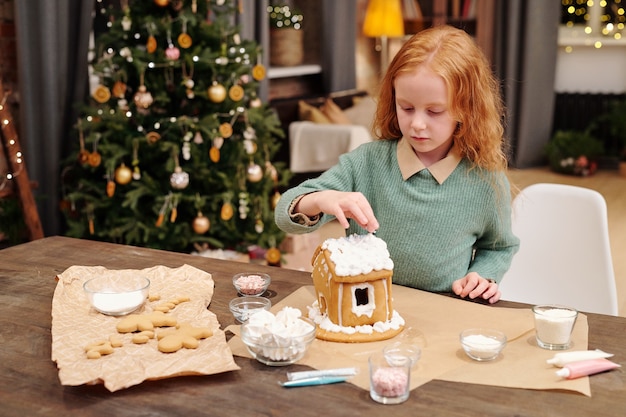 Urocza mała dziewczynka trzymająca rękę nad dachem domku z piernika ozdobiona bitą śmietaną, pomagająca mamie w świątecznym deserze na Boże Narodzenie