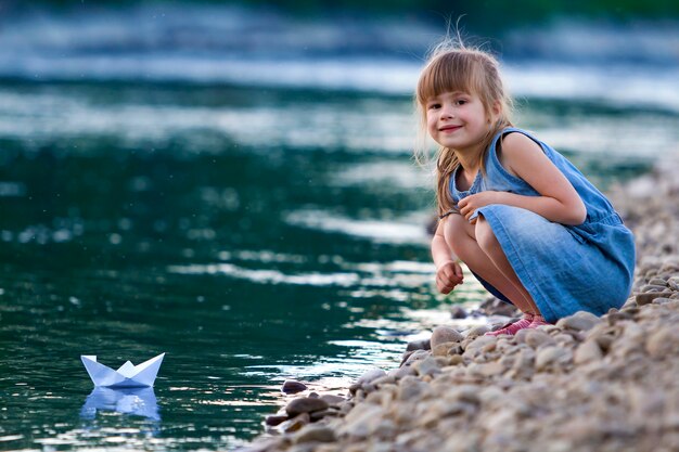 Urocza mała śliczna blond dziewczyna w błękit sukni na otoczaków riverbank bawić się z białego papieru origami łodzią na błękitnej iskrzastej bokeh wodzie. Marzenia i fantazje dotyczące koncepcji szczęśliwego dzieciństwa.