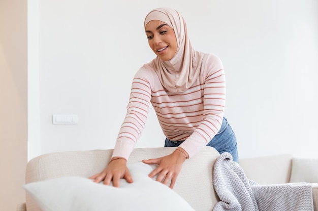 Urocza Młoda Arabska Muzułmanka Kładzie Miękkie Poduszki I Pledy Na Wygodnej Sofie, Dzięki Czemu Jej Dom Jest Przytulny I Ciepły