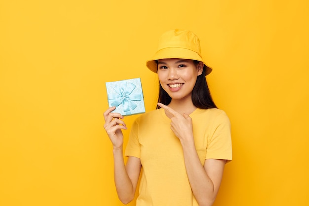 Urocza młoda Azjatka w żółtej koszulce i kapeluszu z prezentem w niezmienionym żółtym tle