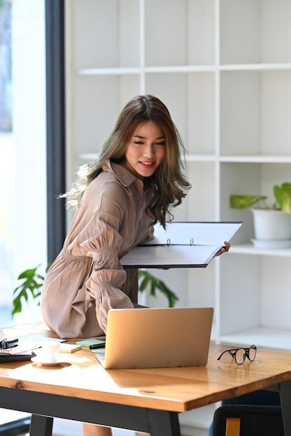 Urocza młoda azjatycka kobieta przedsiębiorca siedzi na biurku i korzysta z laptopa
