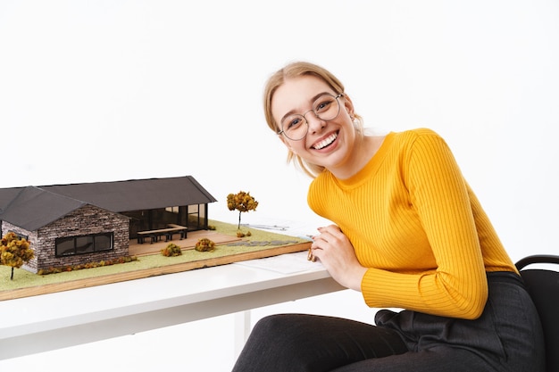Urocza Młoda Kobieta Architekt Siedząca Przy Biurku Z Modelem Domu Na Białym Tle Nad Białą ścianą