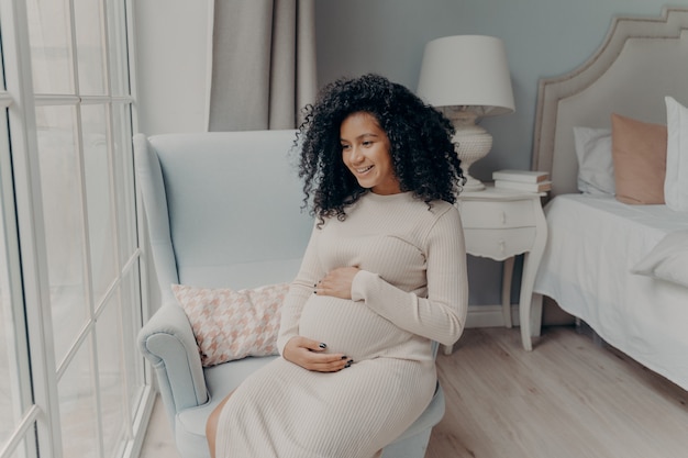 Urocza młoda kobieta w ciąży mieszanej rasy, przyszła mama wygląda przez duże okno i marzy o macierzyństwie siedząc bokiem na fotelu w sypialni, trzymając się za brzuch obiema rękami i uśmiechając się