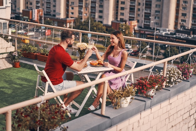 Urocza młoda para w swobodnych ubraniach, wznosząca toast i uśmiechająca się, siedząc na patio na dachu na świeżym powietrzu