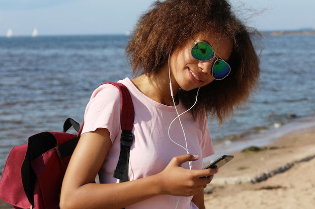 Urocza niesamowita afroamerykańska młoda kobieta w okularach przeciwsłonecznych słuchająca muzyki w słuchawkach na swoim telefonie komórkowym