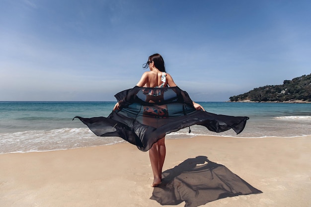 Urocza niesamowita seksowna modelka w bikini i czarnym szlafroku spaceruje po morskiej plaży, a jej szlafrok rozwija się na wietrze. Widok z tyłu. Koncepcja wakacji.
