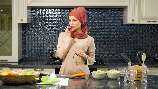 Urocza orientalna kobieta w hidżabie i długiej sukience stoi przy kuchennym stole i zjada warzywa, przygotowując jedzenie na rodzinne zbliżenie